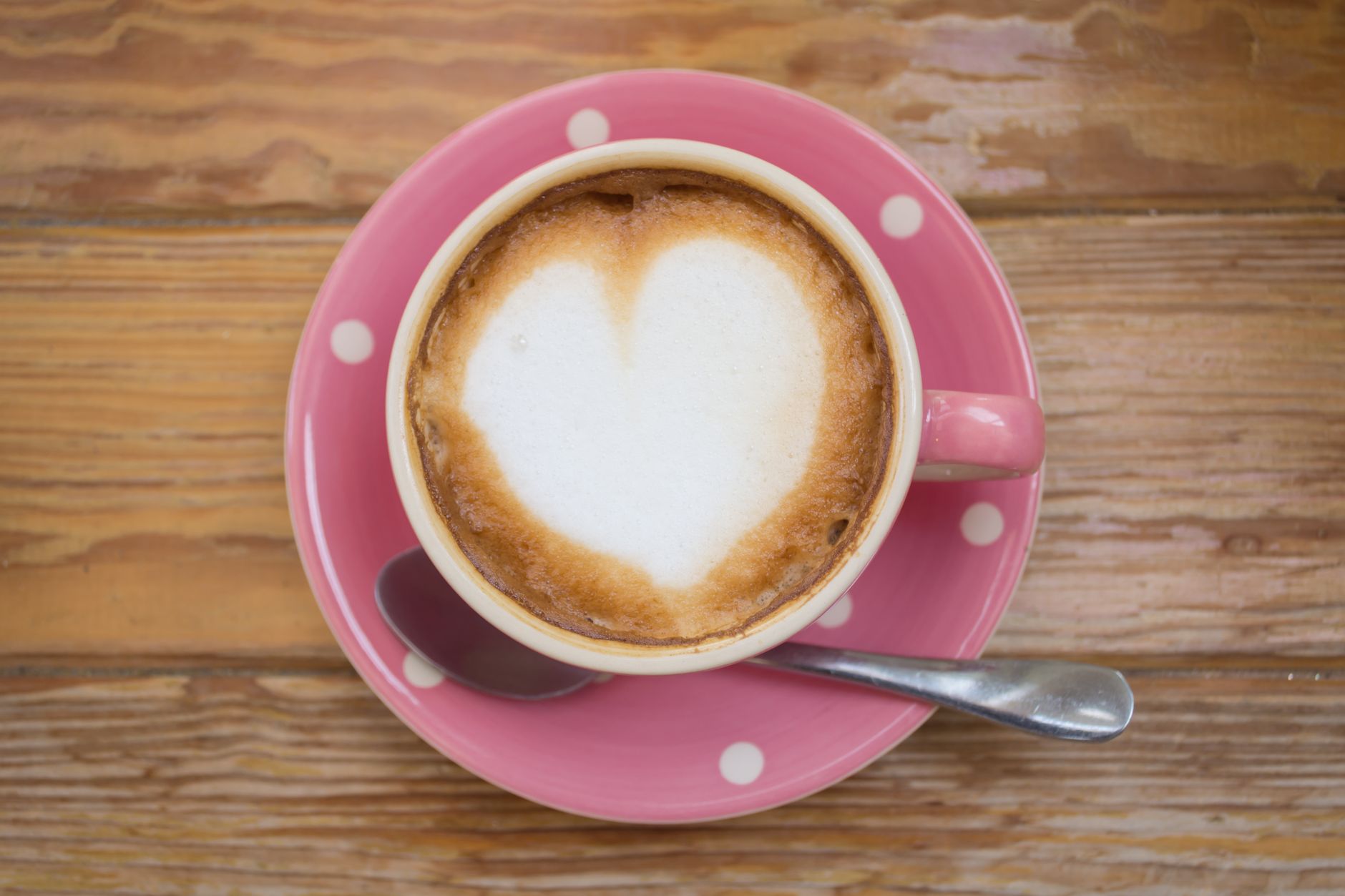 A top view of heart-shaped latte art in a pink polka dot cup on a wooden table.
