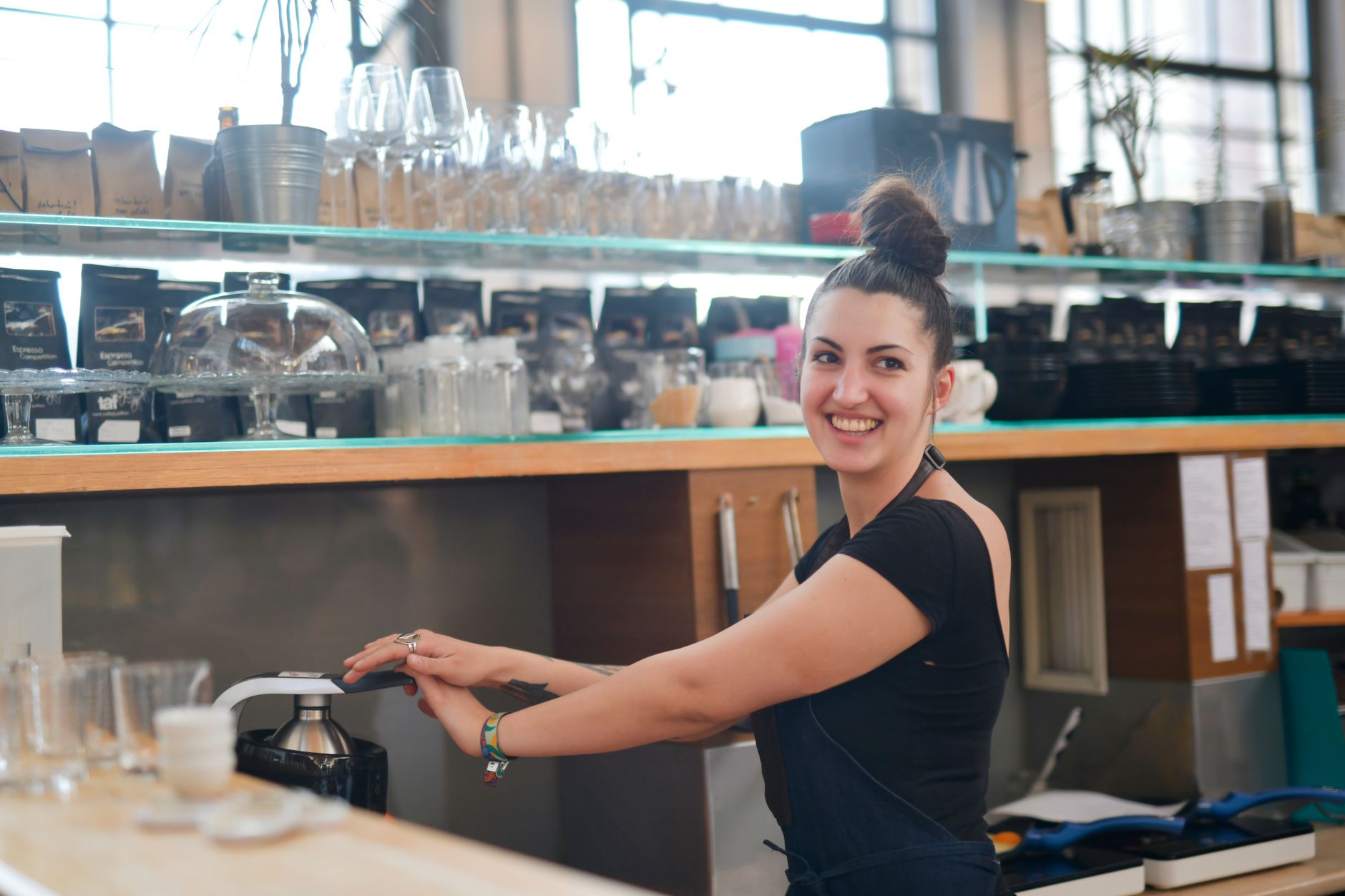 Positive young female bartender in black apron making fresh fruit drink in juicer while working in modern cafe and looking away