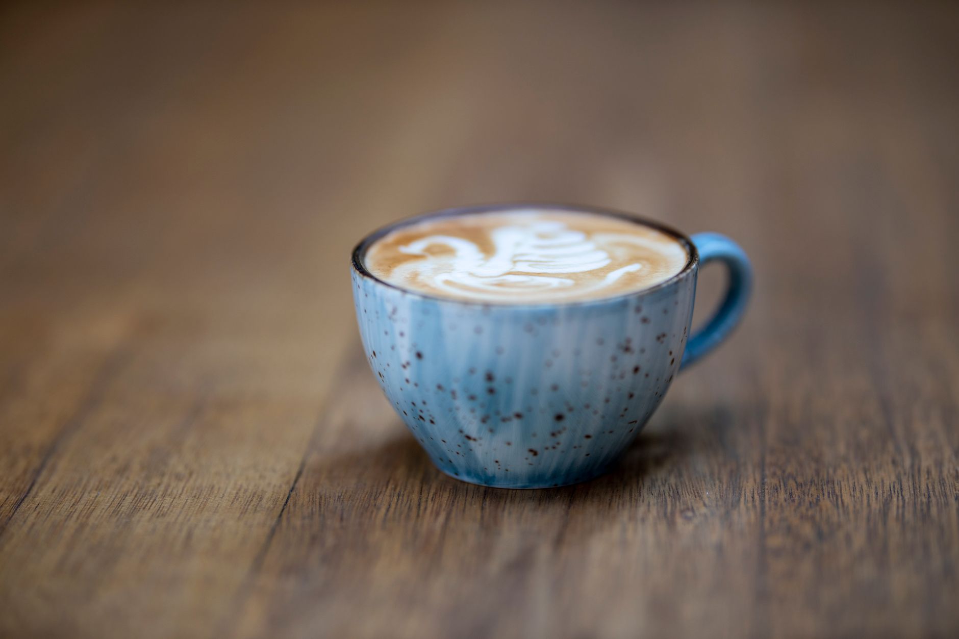 A cappuccino with intricate latte art served in a blue speckled cup on a wooden table.