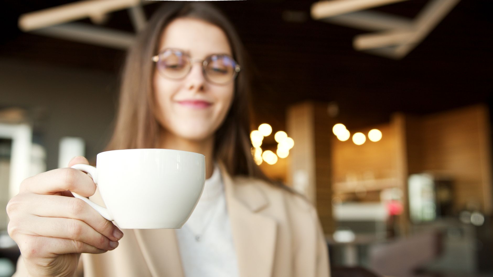A woman with glasses smiles while holding a cup of coffee inside a cozy cafe.