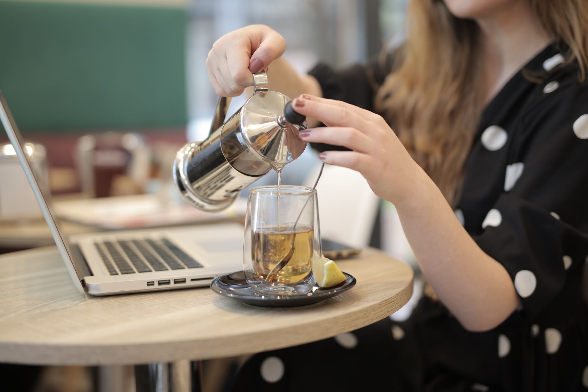 Side view of crop female in polka dot dress sitting at table with laptop in cafeteria and pouring hot drink into transparent glass mug with double bottom