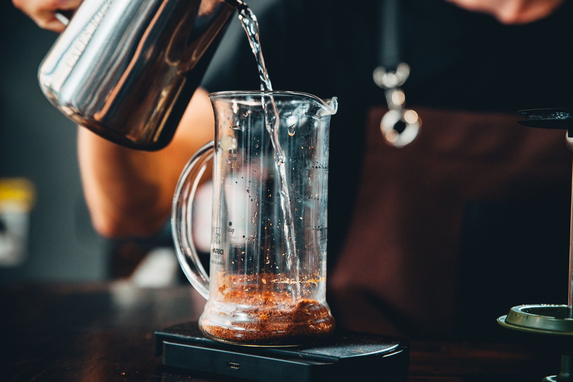 Hands pouring hot water into glass pitcher with coffee grounds for brewing.
