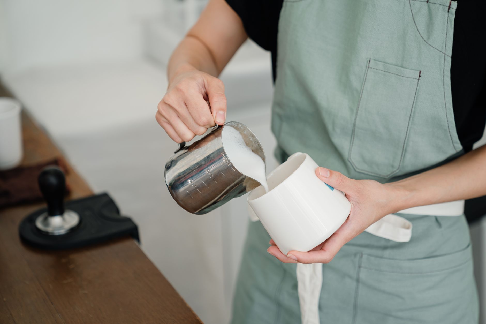 From above of crop young anonymous person in apron pouring milk into white mug while preparing aromatic latte