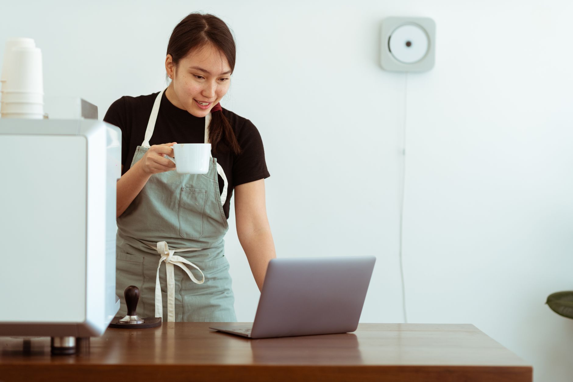 Positive young Asian female in apron with white cup of hot beverage working on modern netbook in light kitchen against white wall