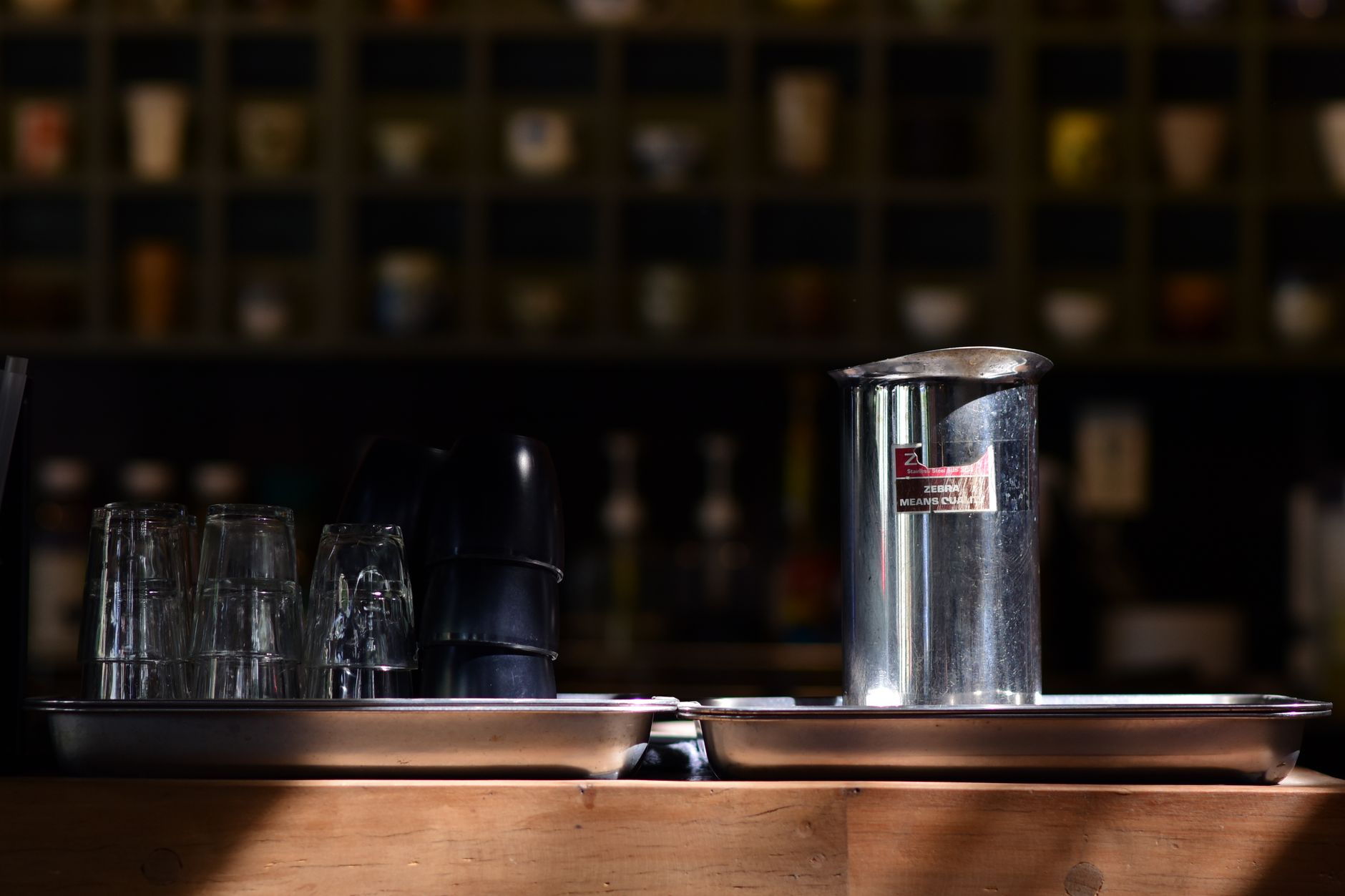 Elegant arrangement of glassware and a metal pitcher on a wooden counter in a dimly lit cafe setting.