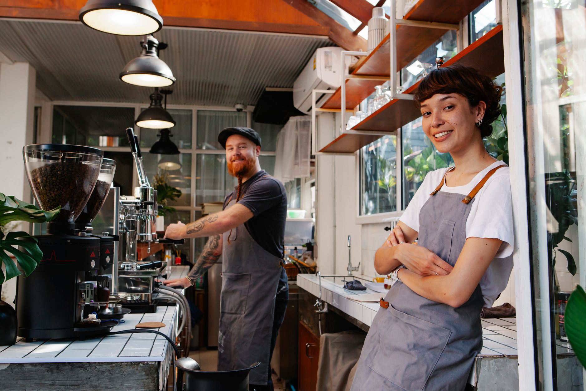 Cheerful young baristas in aprons working together in contemporary cafeteria kitchen and looking at camera with smiles