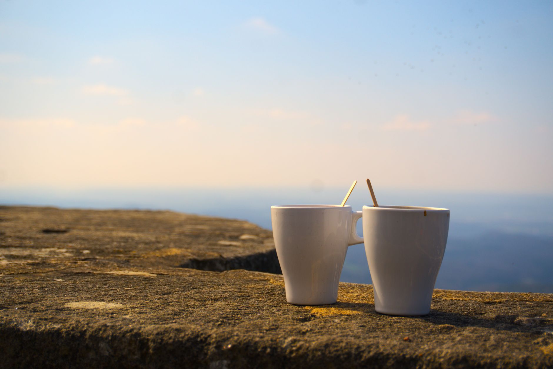 Two white coffee mugs on a stone wall with a scenic sunrise view.