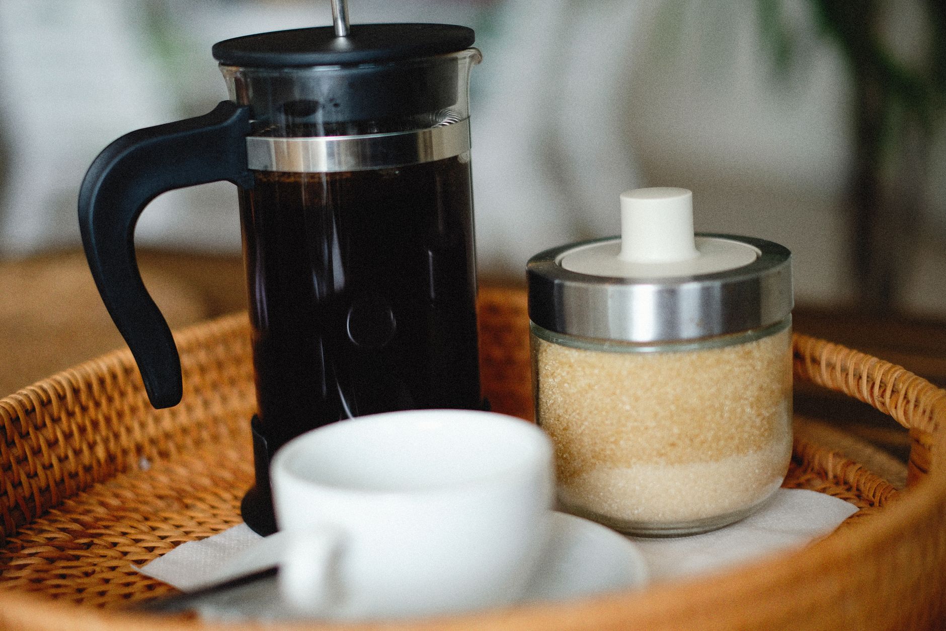 Close-up of a French press, sugar jar, and coffee cup on a wicker tray.
