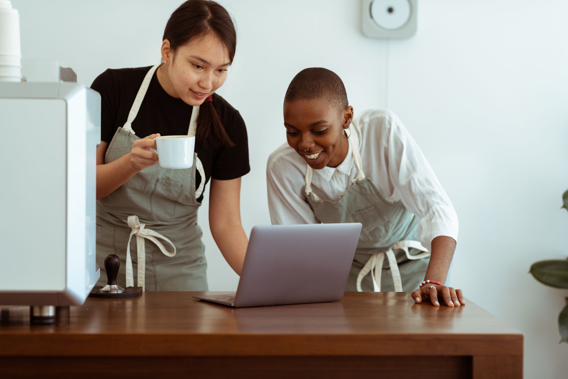 Cheerful smiling young diverse female waitresses in aprons with cup of hot beverage browsing netbook while standing near wooden table in light kitchen