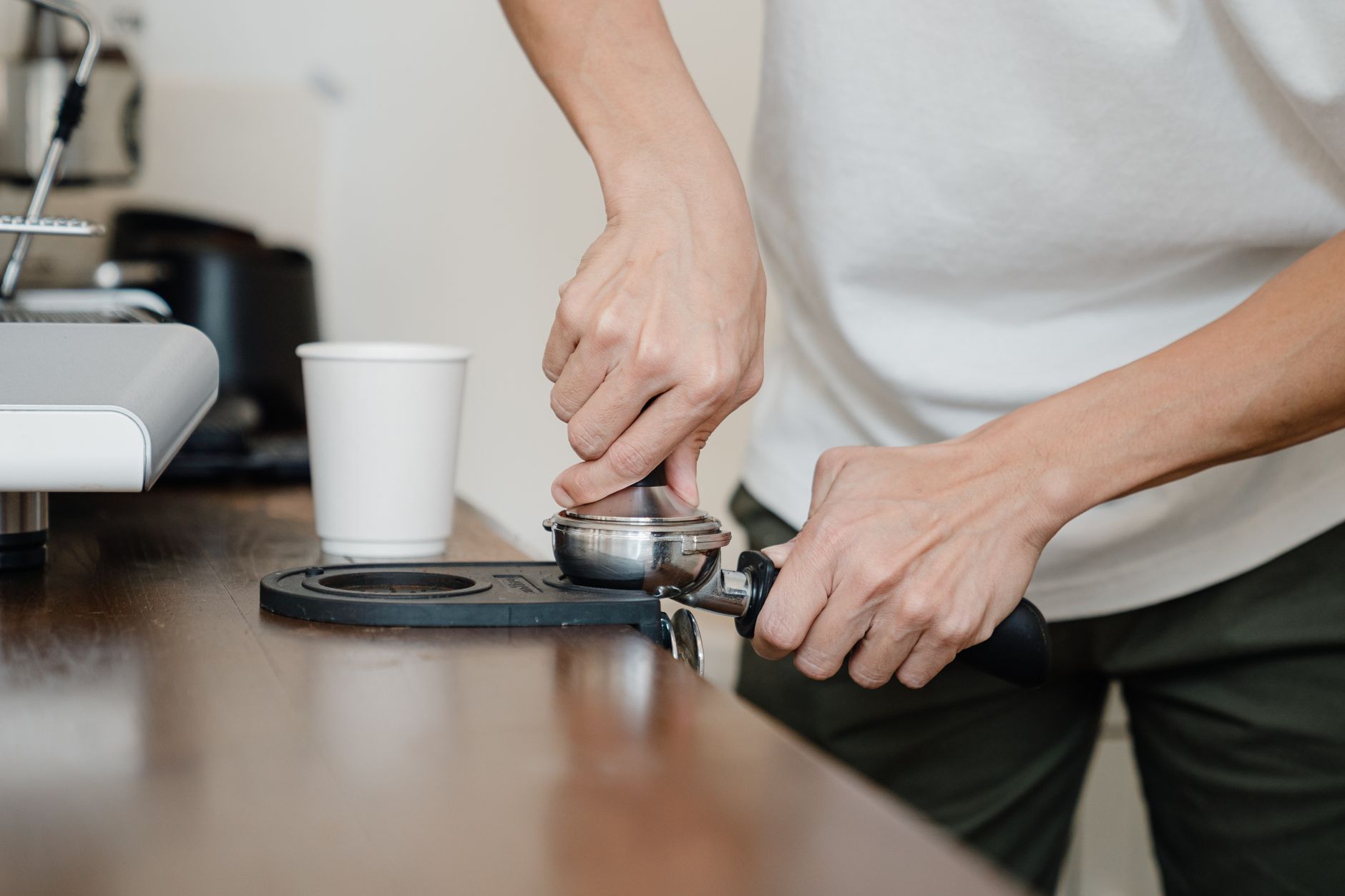 Crop anonymous male barista in casual wear manually pressing coffee beans into metal holder by using tamper while standing nearby professional coffee machine and white cup