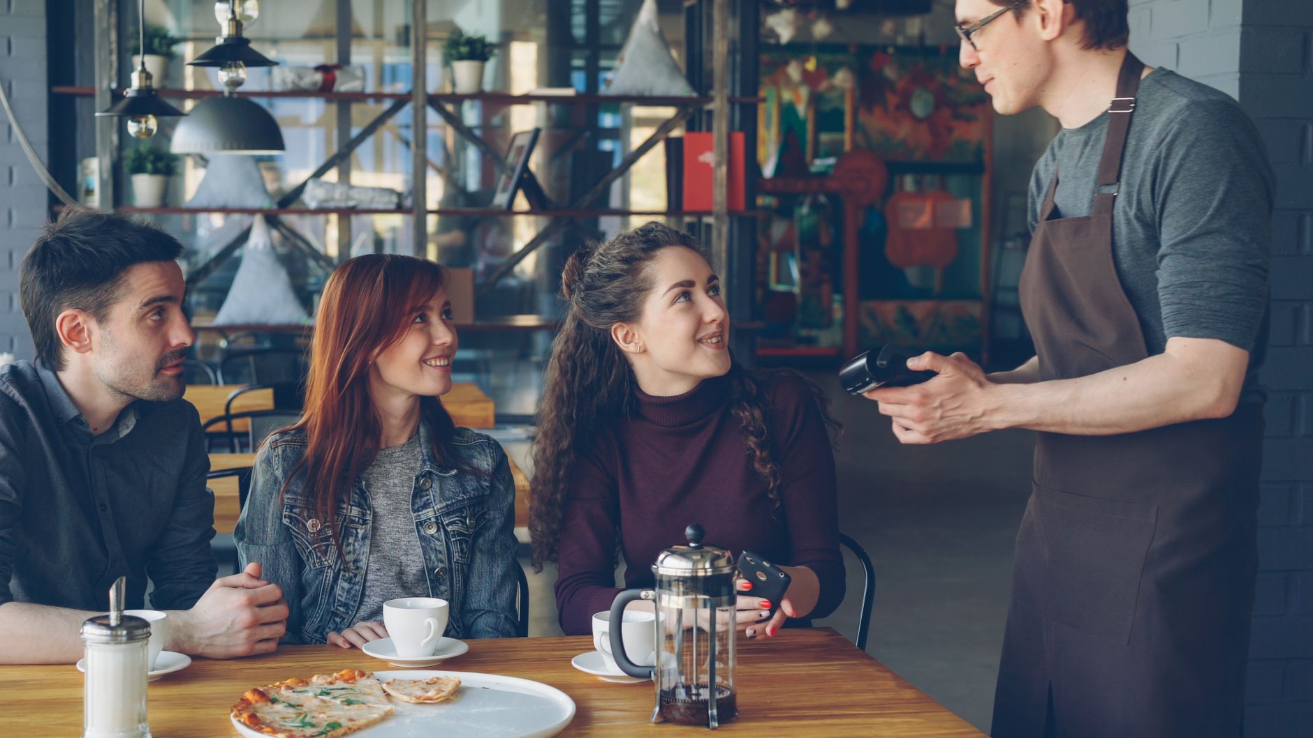 Three friends interact with a waiter in a cozy café, creating a warm and inviting scene.