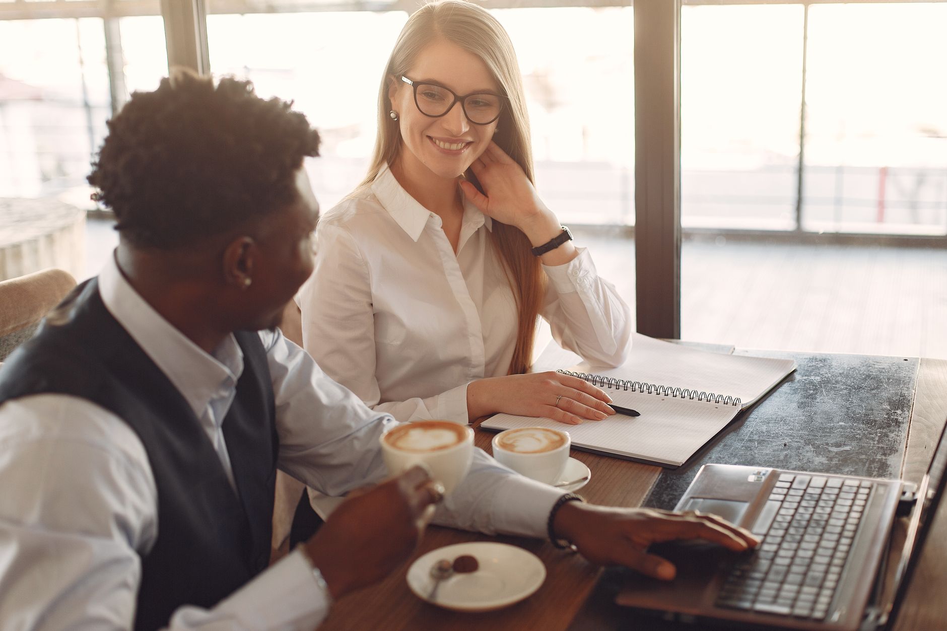 Side view of positive young multiethnic coworkers in formal clothes sitting at wooden table and drinking coffee while working together in modern workspace
