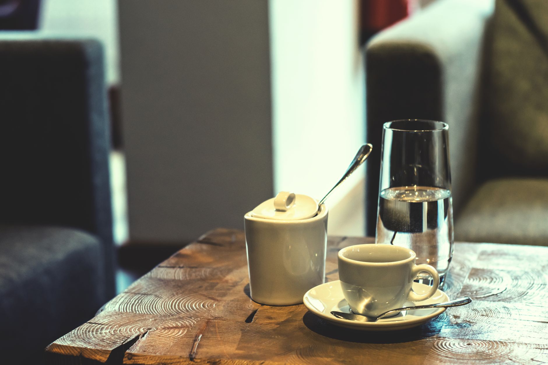 A warm setting featuring an espresso cup, sugar pot, and water glass on a wooden table.
