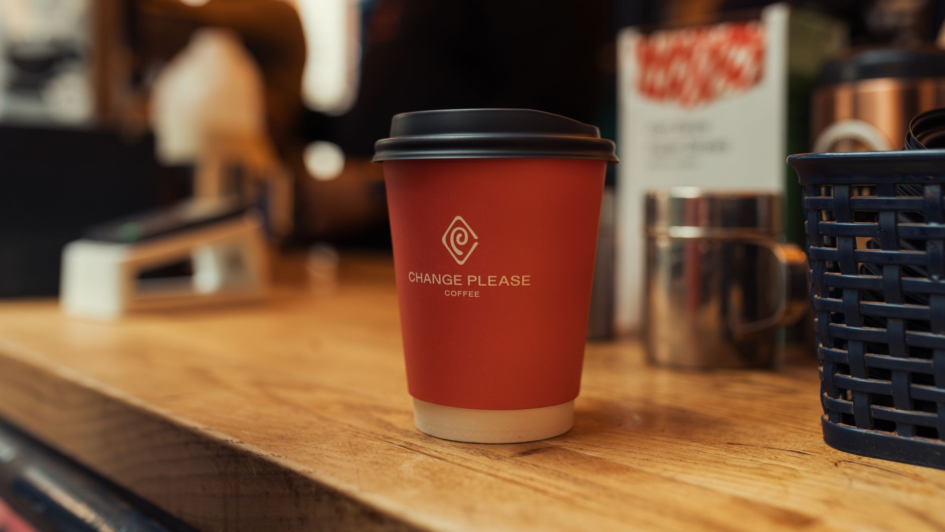 A red coffee cup with the logo 'Change Please' on a wooden counter in a cozy indoor setting.
