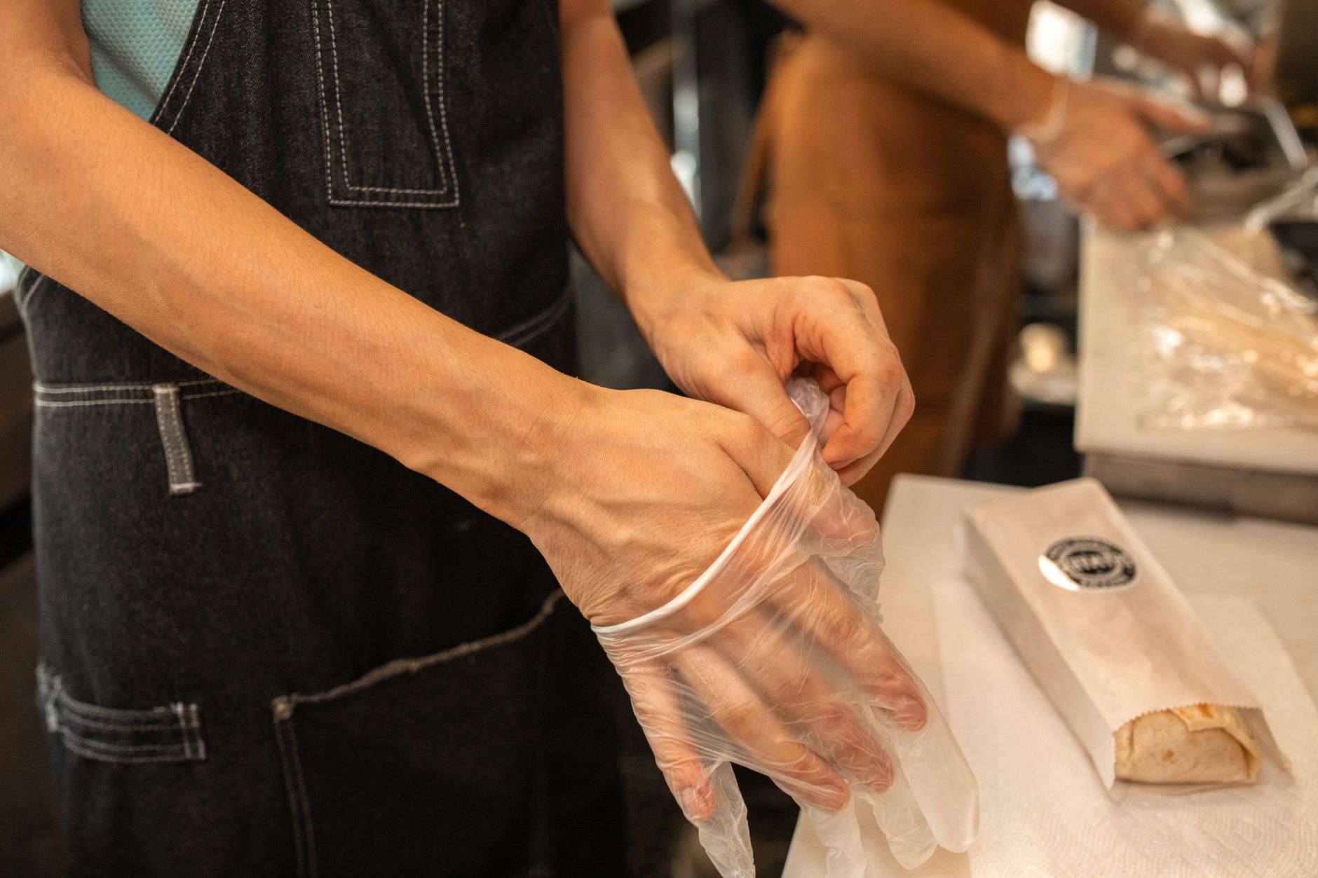 A person wearing an apron puts on plastic gloves in a kitchen environment, preparing for food handling.