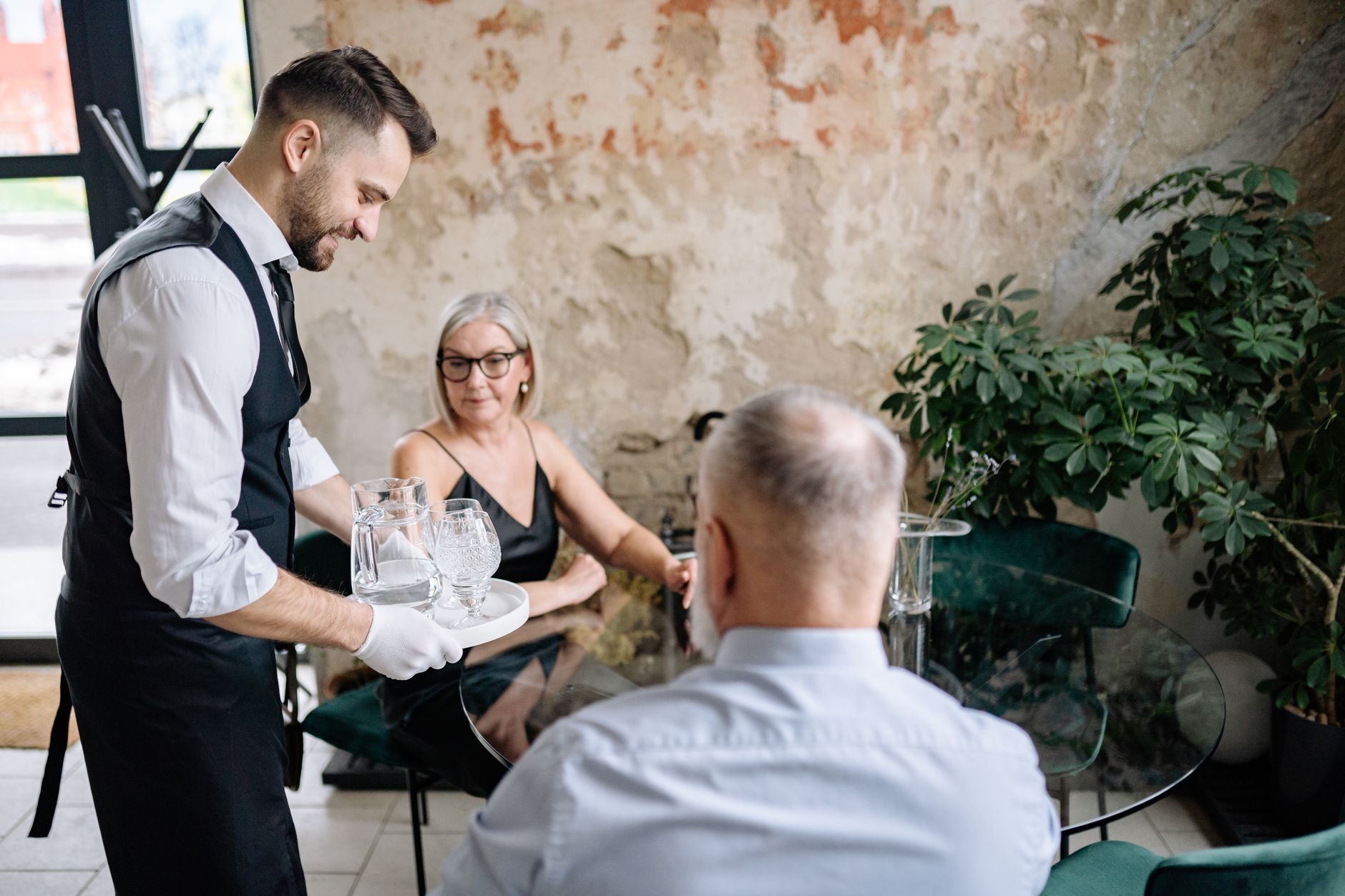 A waiter serving drinks to a senior couple in a stylish, rustic restaurant setting.