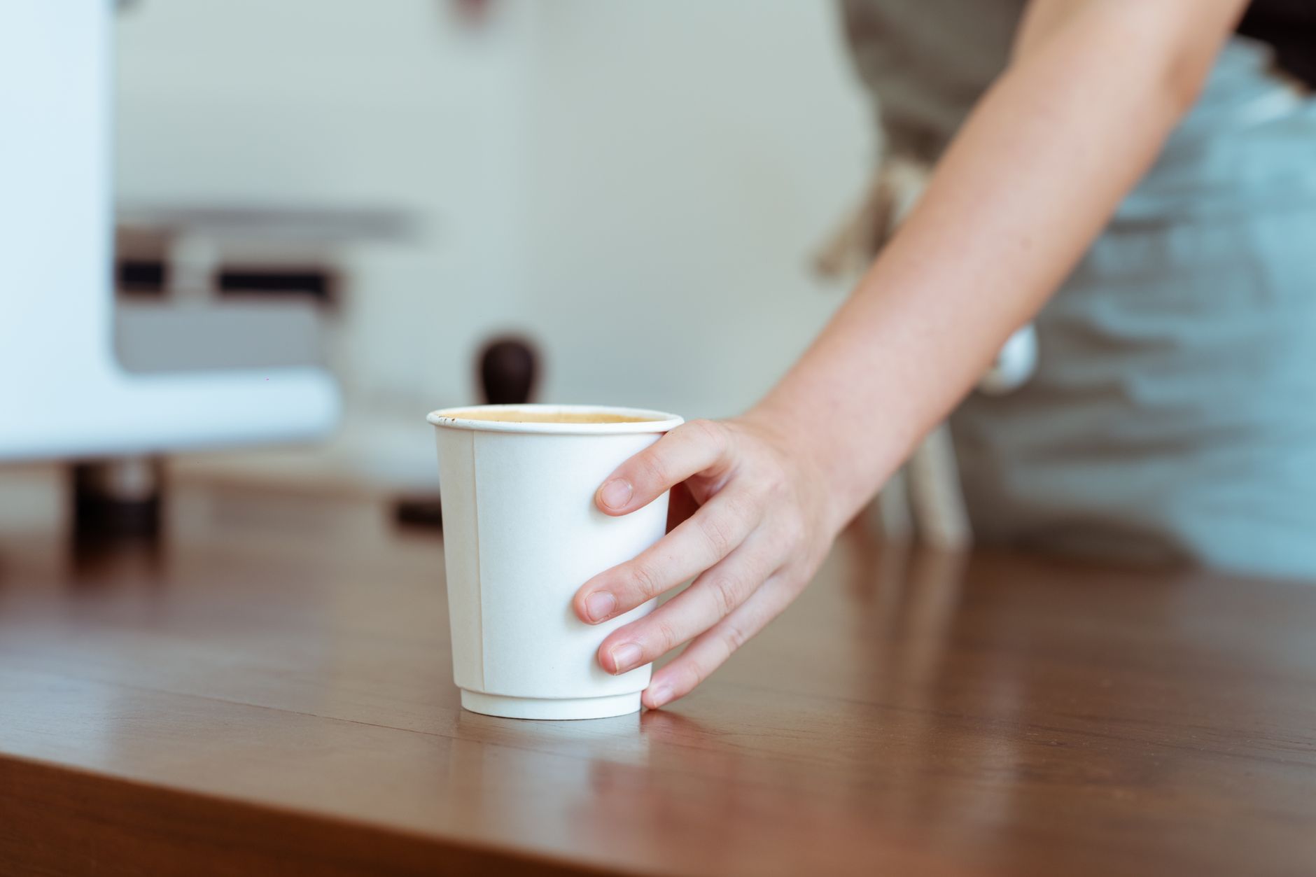 Crop unrecognizable female barista in apron passing white paper cup with aromatic coffee while standing nearby professional coffee machine in contemporary cafe