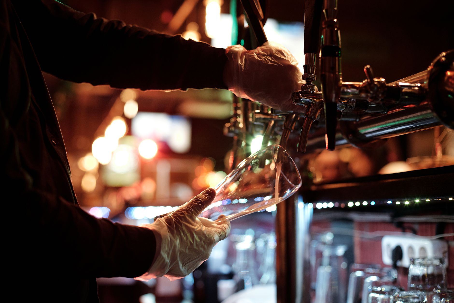 Bartender with gloves pouring beer into a glass in a dimly lit nightclub.