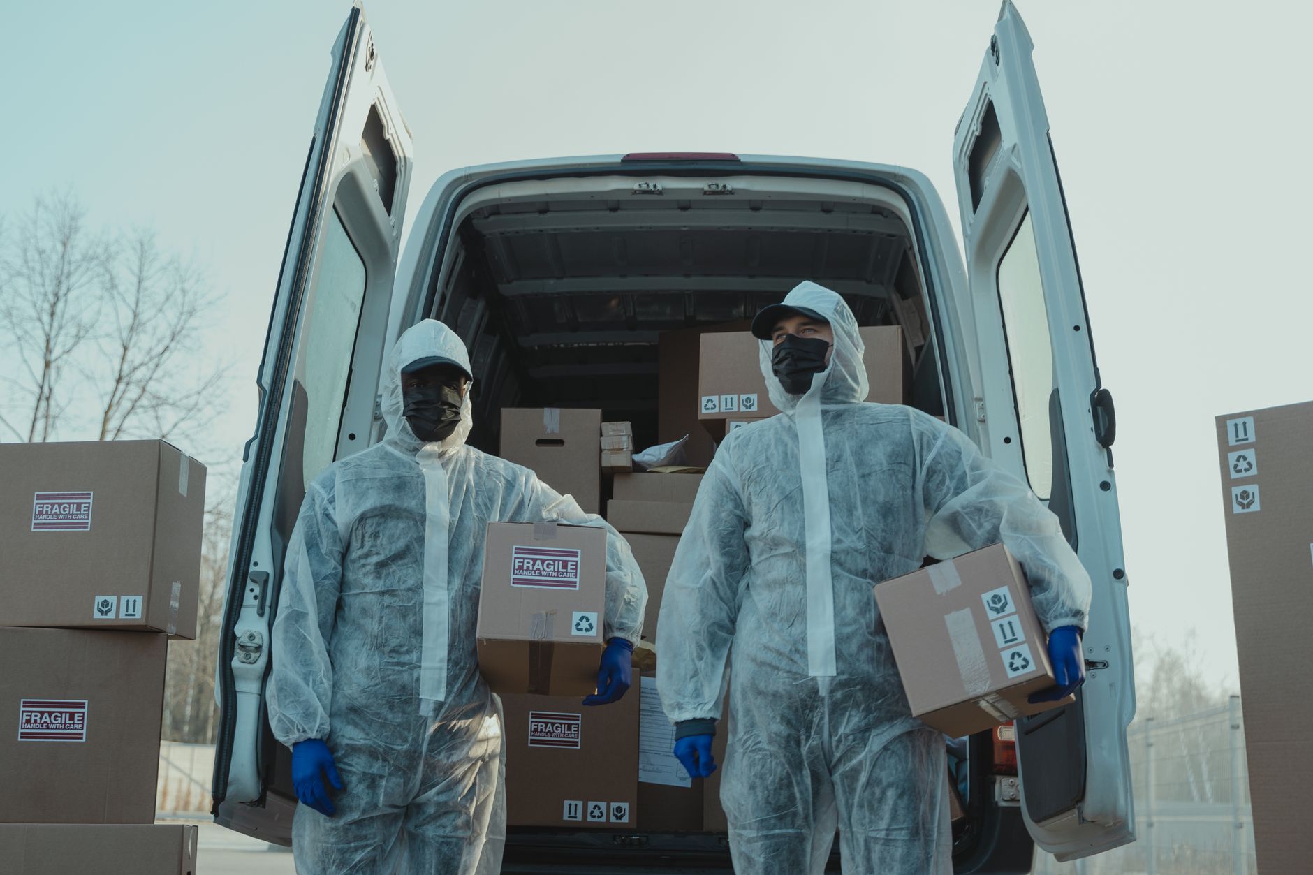 Two workers in PPE gear handling packages by a delivery van during daytime.