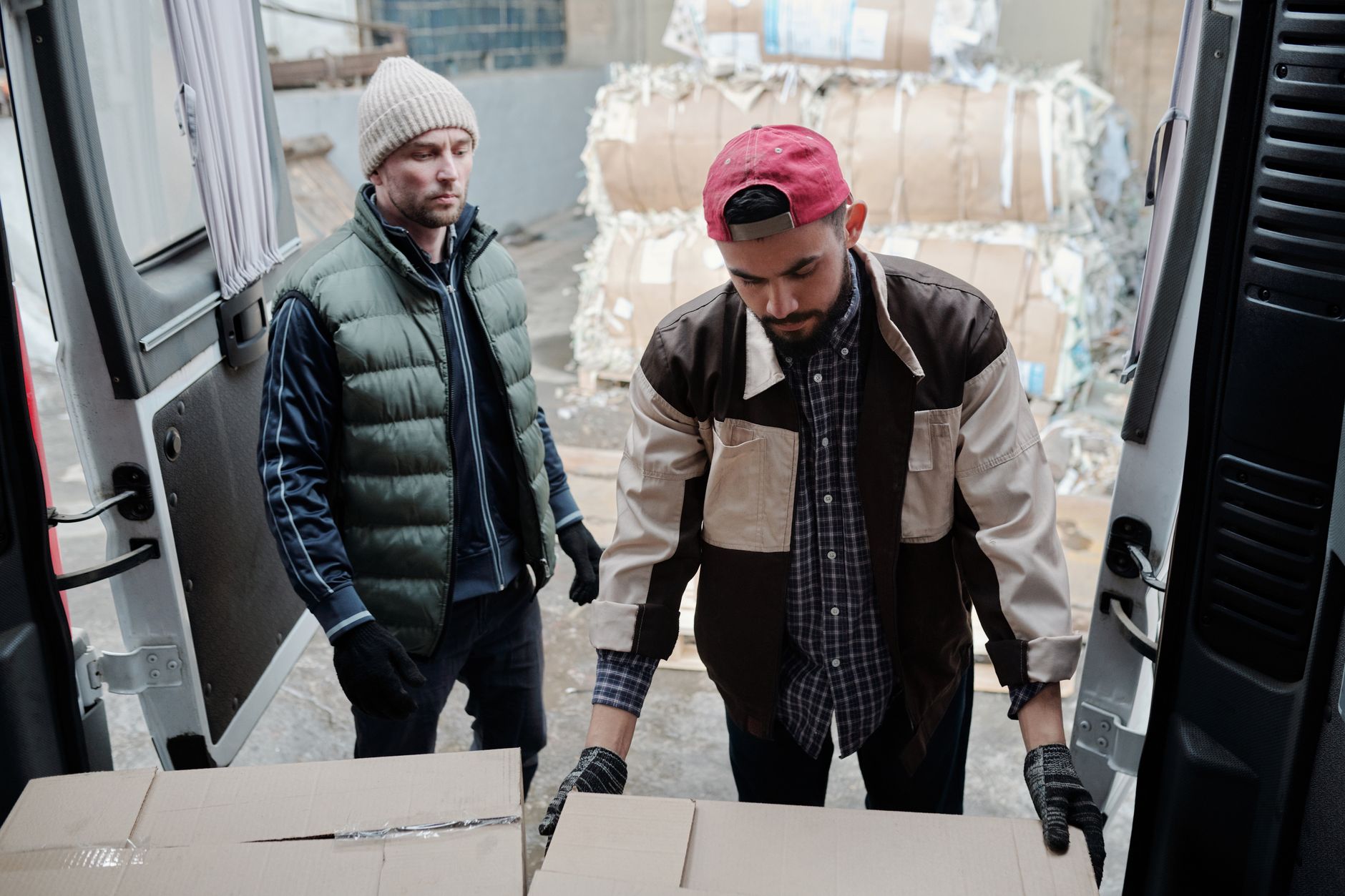 Two men loading cardboard boxes into a delivery van at an urban site.
