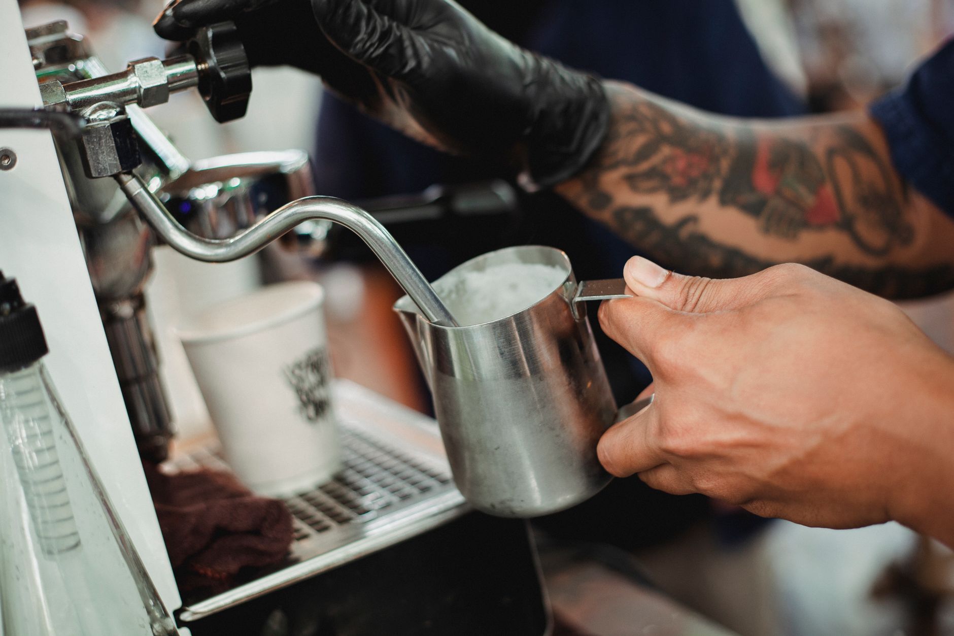Crop unrecognizable tattooed male barista in black glove frothing warm milk in pitcher with coffee machine steam wand while working in modern cafe