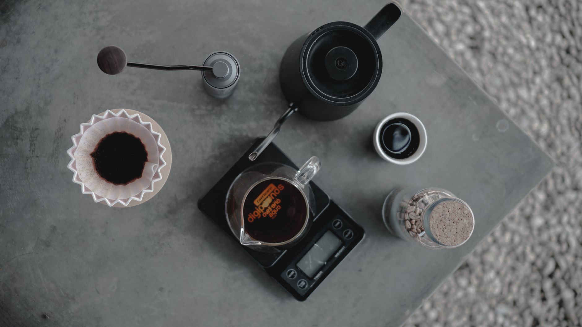 Aerial view of coffee brewing setup with pour-over, grinder, and scale on a textured surface.