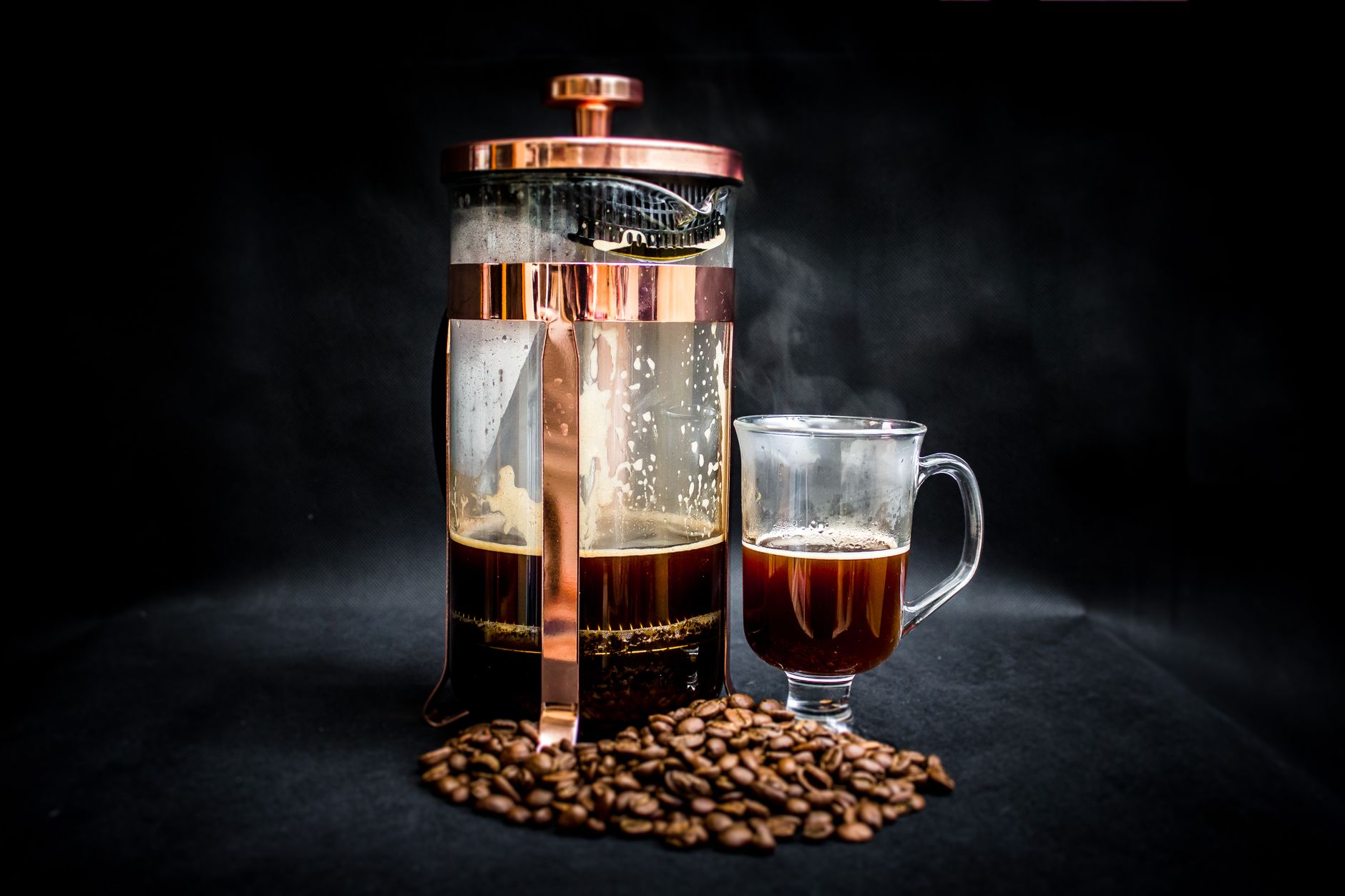 Artistic shot of a French press and glass mug with hot coffee and scattered beans.