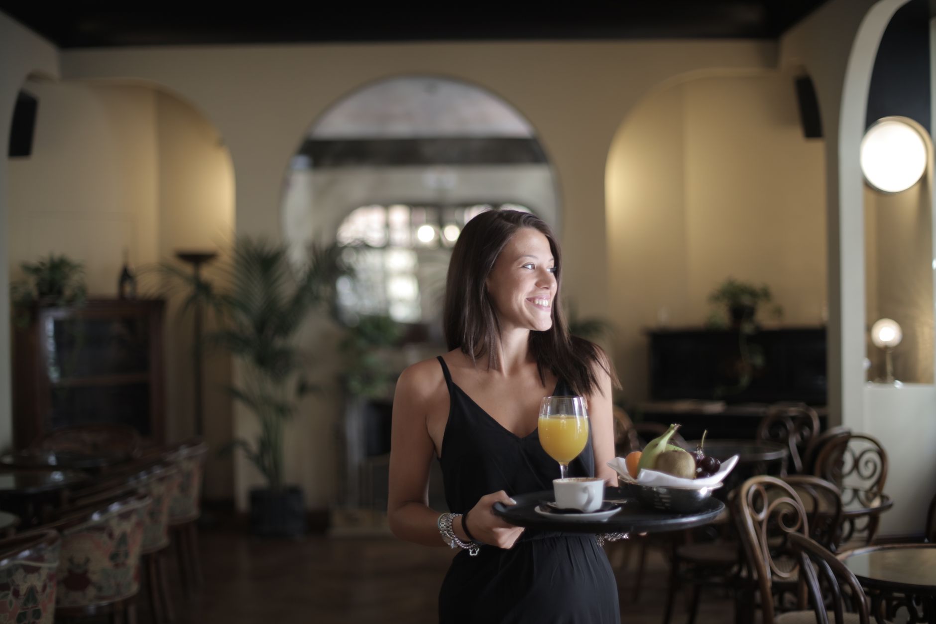 Cheerful young woman in elegant black dress holding tray with healthy breakfast with fruits and juice while standing in cozy cafe