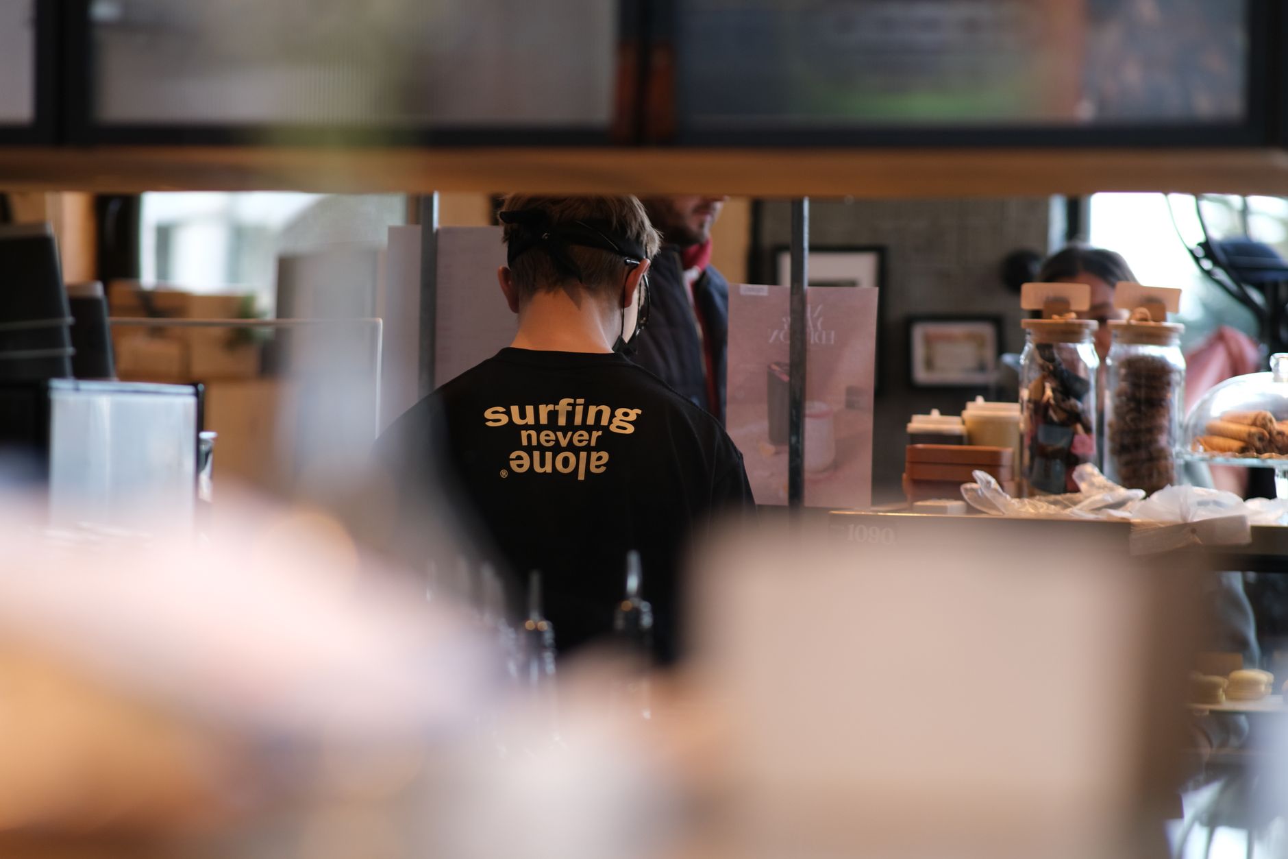Barista working behind a counter in a lively cafe, wearing a shirt with text.