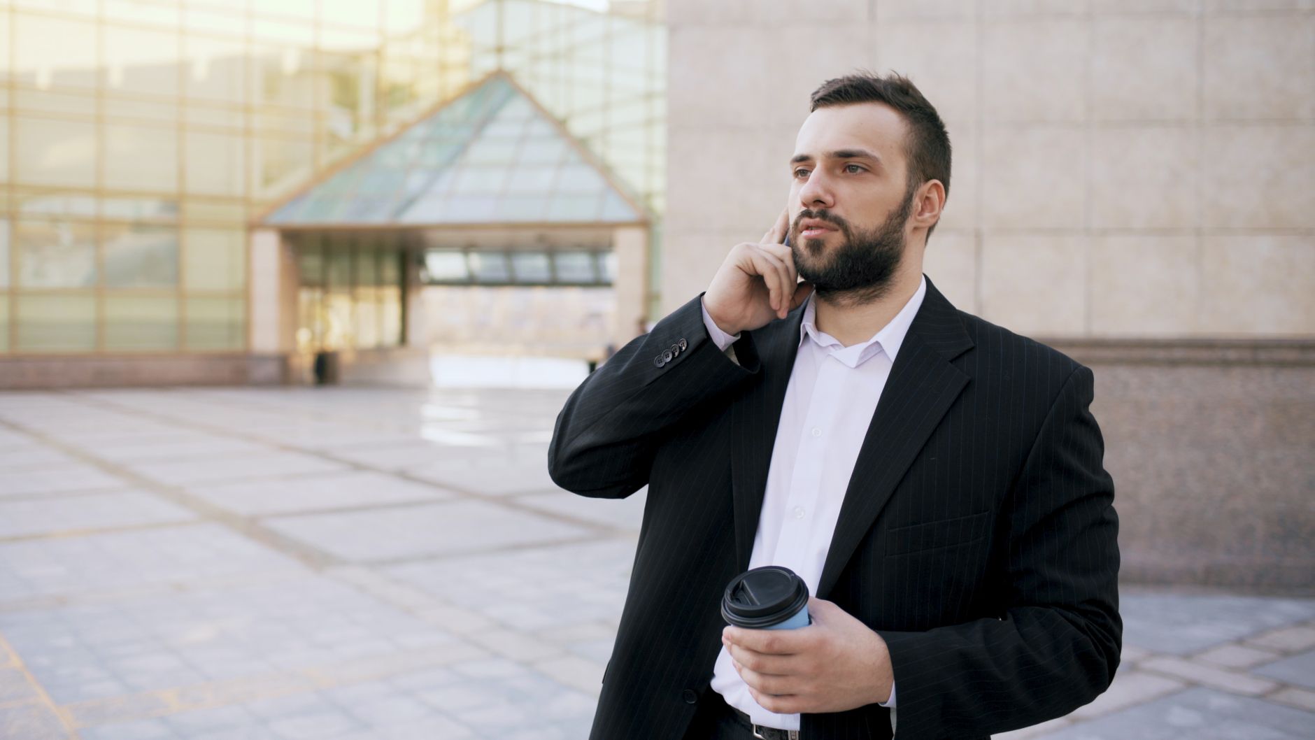 A professional businessman with a coffee cup makes a phone call outside a modern office building.