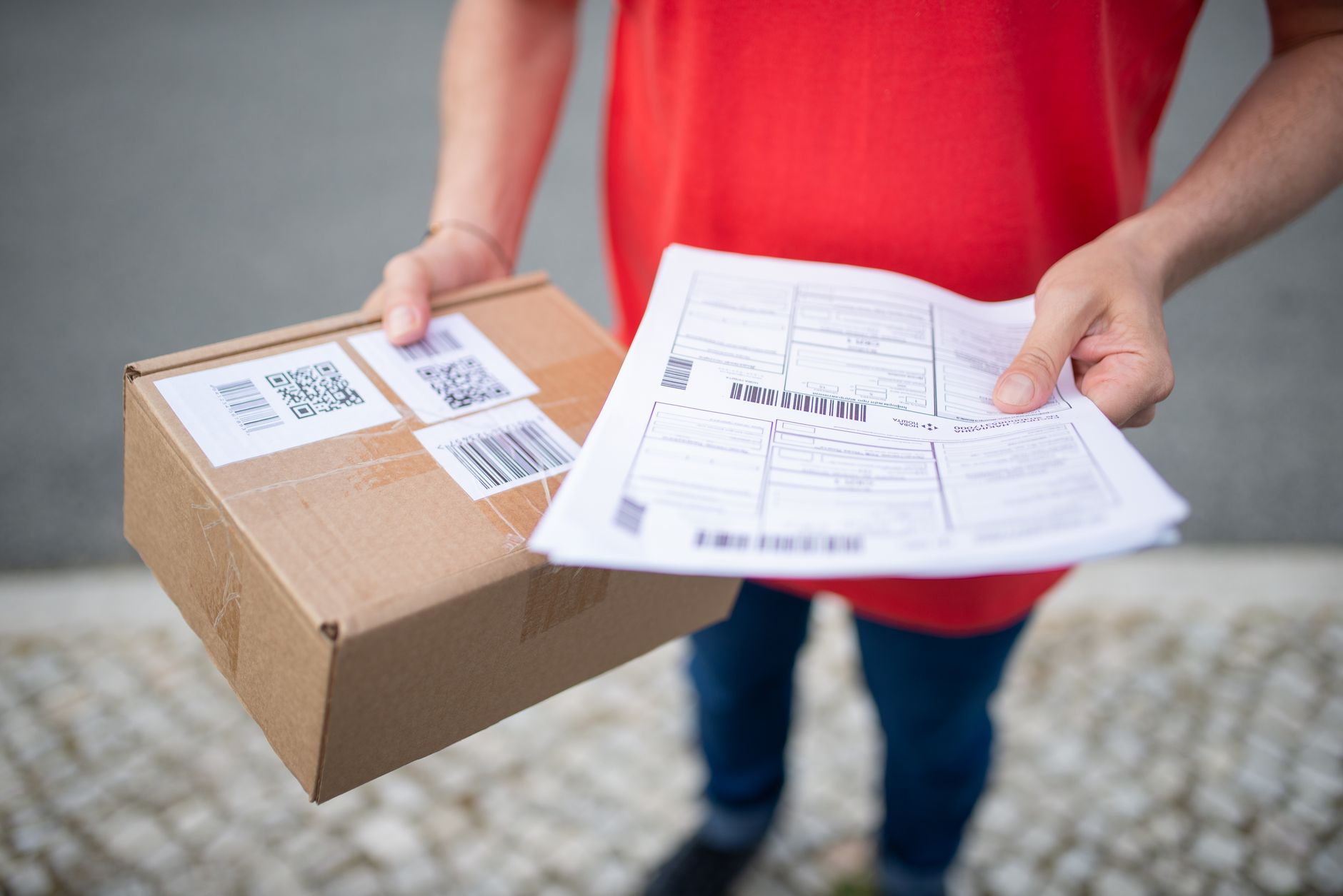 Close-up of person in red shirt holding a brown package and delivery documents outside.