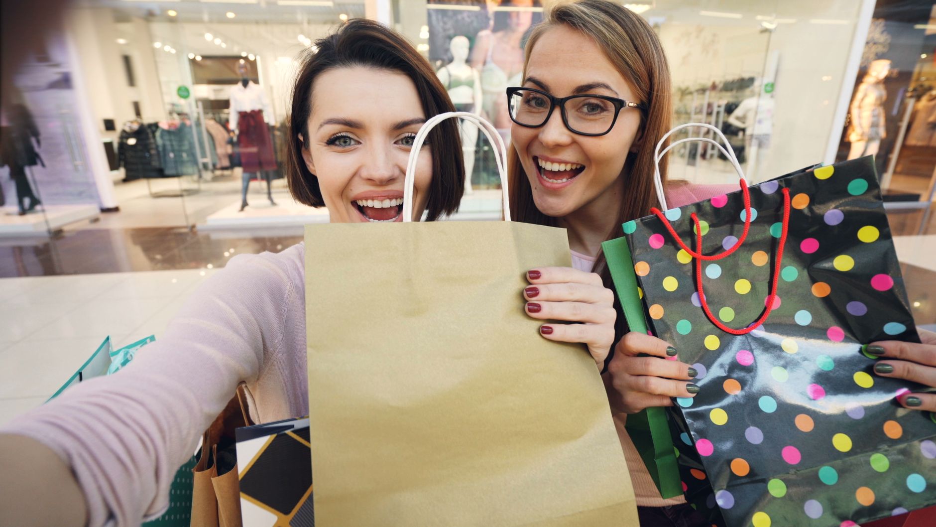 Two women smiling with shopping bags in a mall environment.