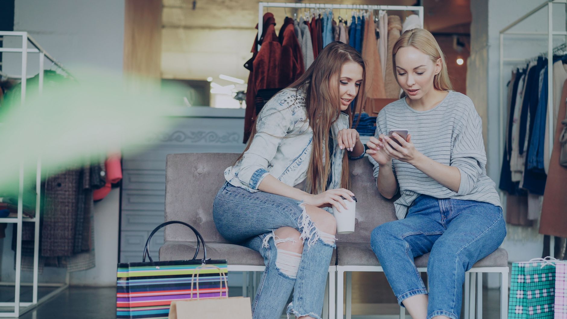 Two women sitting in a boutique, sharing a moment on a smartphone. Retail shopping and friendship.