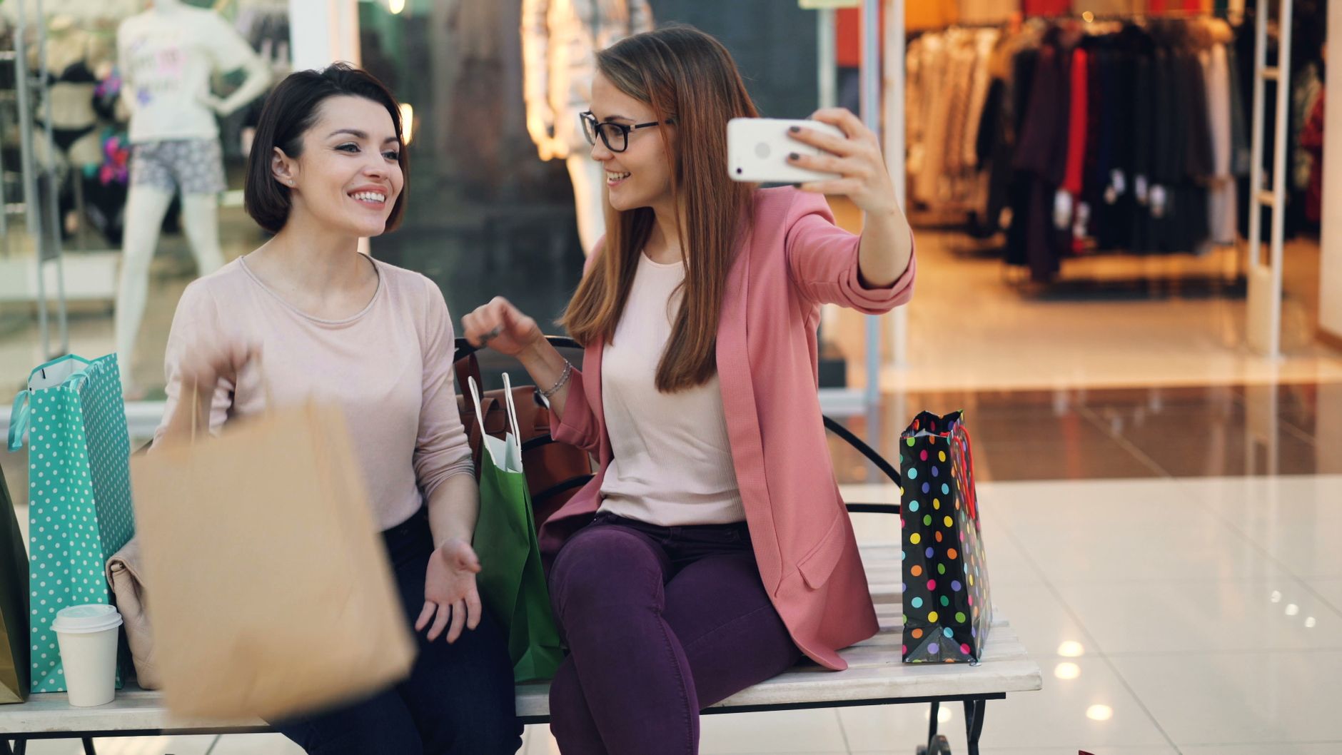 Two women happily taking a selfie with shopping bags in a mall.