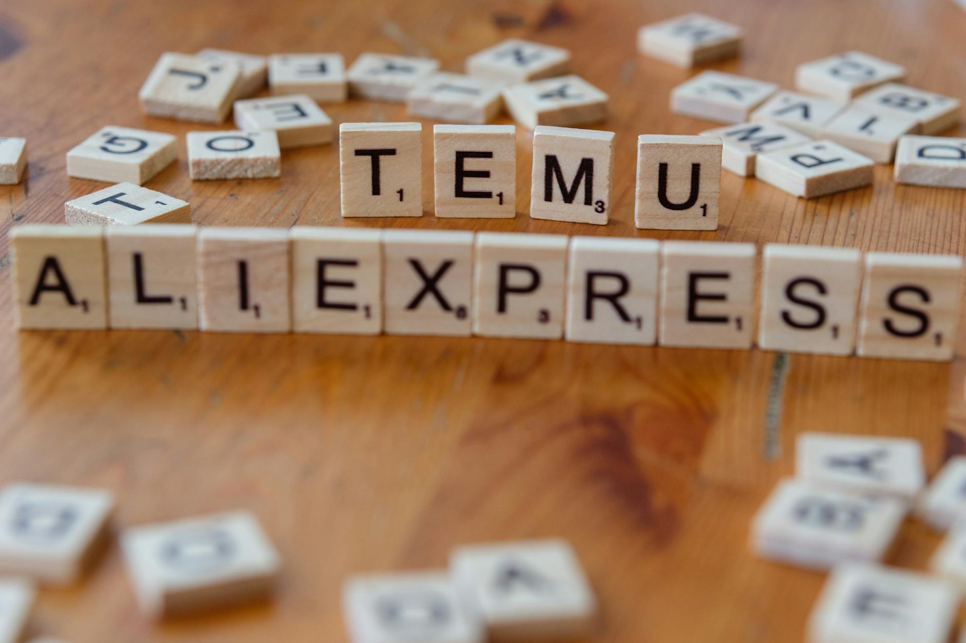 Close-up of Scrabble tiles on a wooden surface spelling 'TEMU' and 'ALIEXPRESS' amid scattered letters.