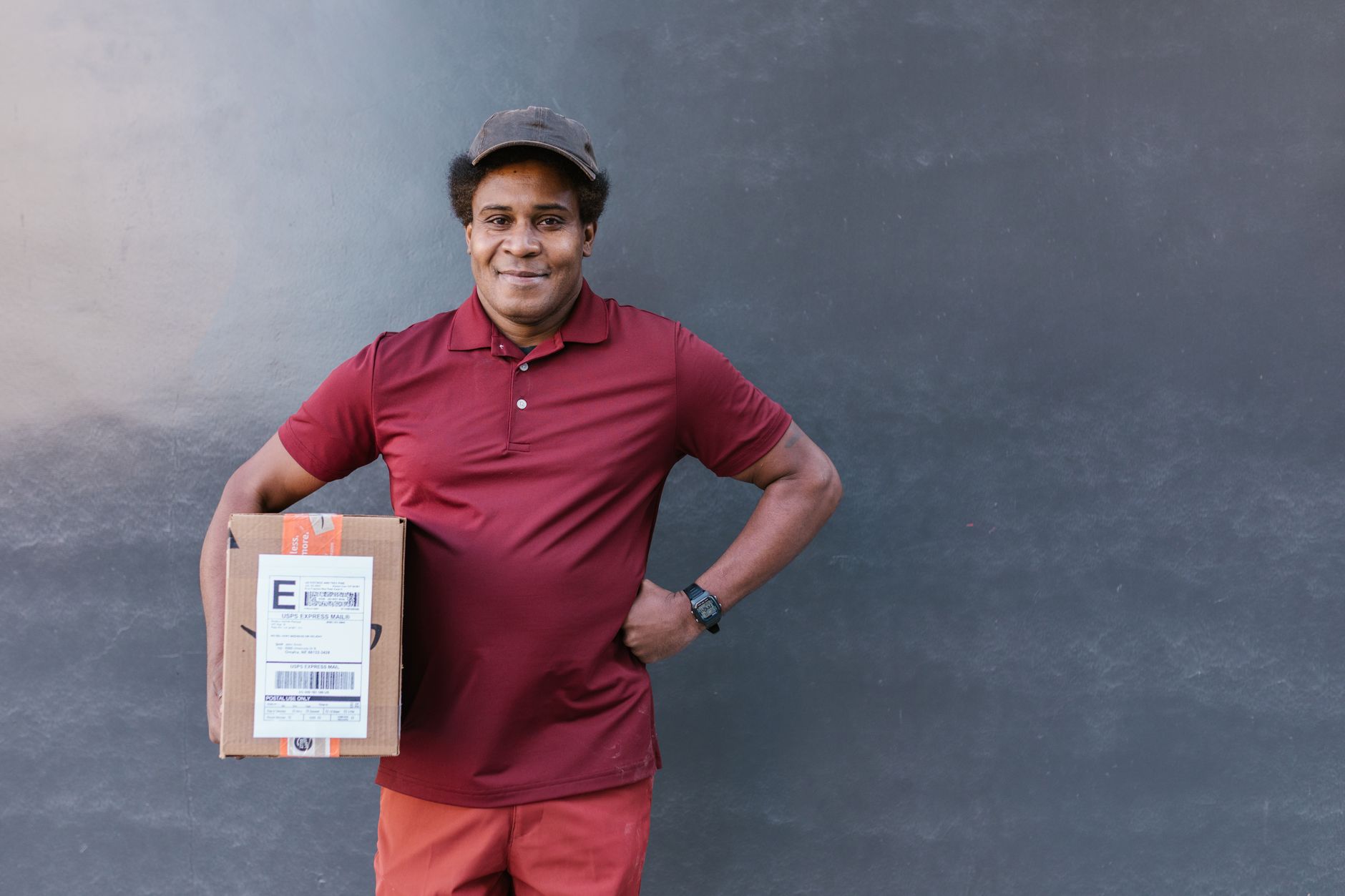Smiling delivery man in red uniform holding a package against a gray wall outdoors.