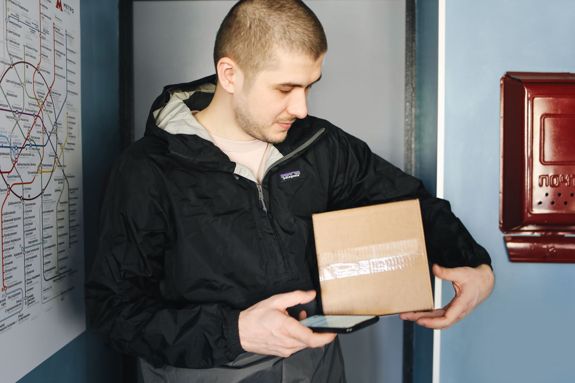 A delivery man holds a package and uses his smartphone inside a building.