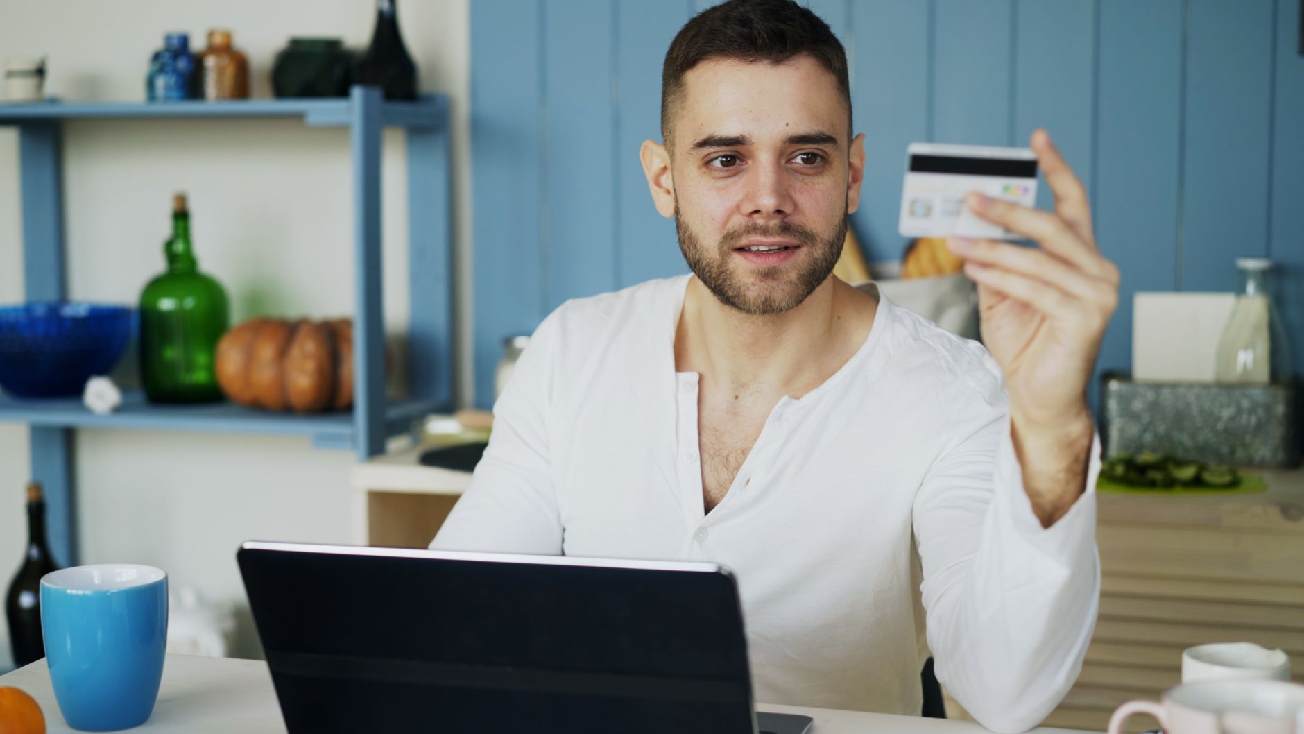 A man in a white shirt sits at a desk, using a laptop and holding a credit card.