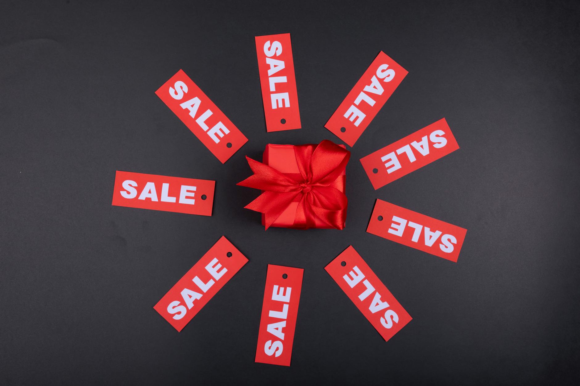 Red gift box with ribbon surrounded by sale tags on a black background.