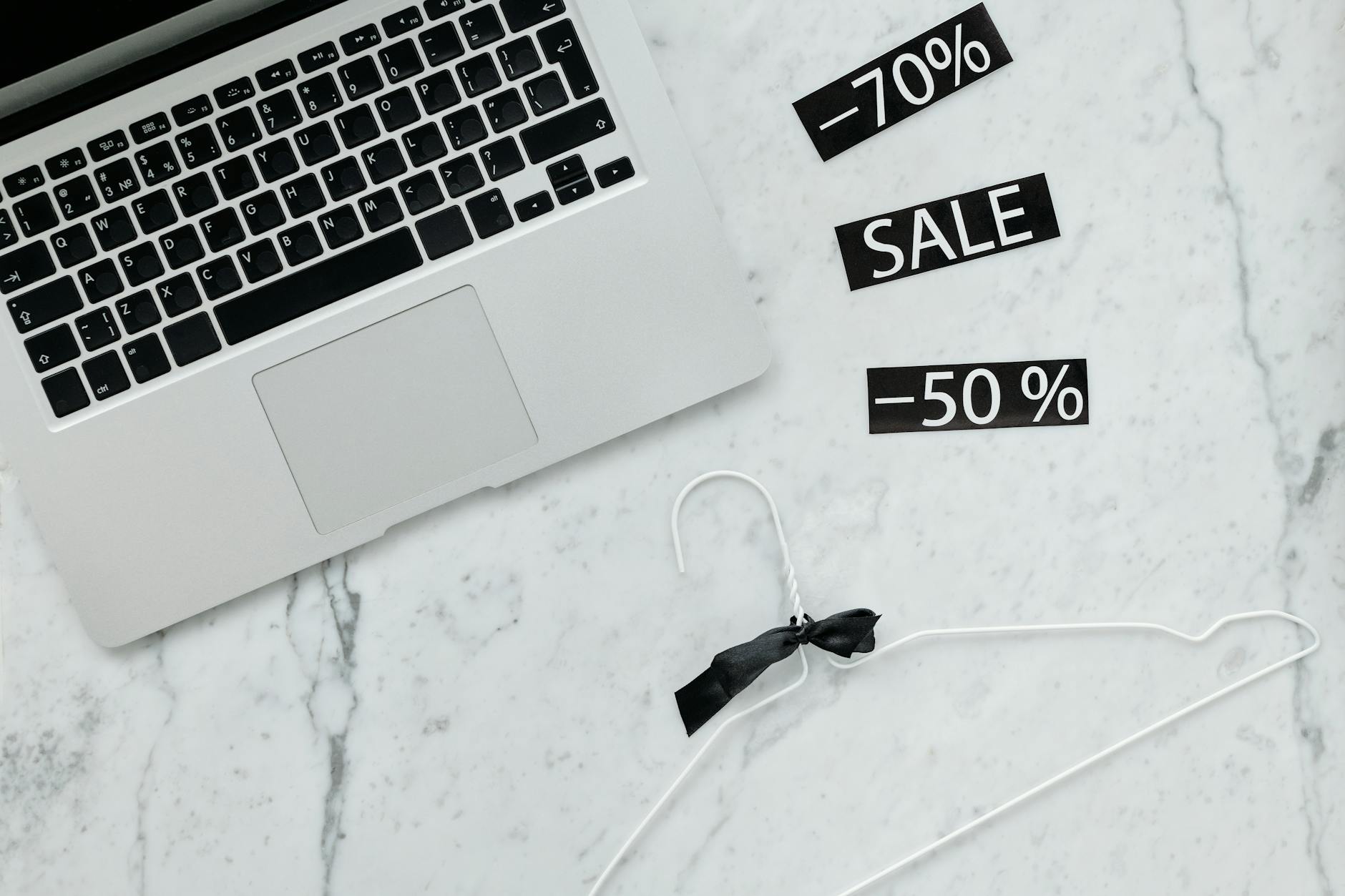 Flat lay of a laptop, hanger, and sale stickers on a marble desk, ideal for Black Friday themes.