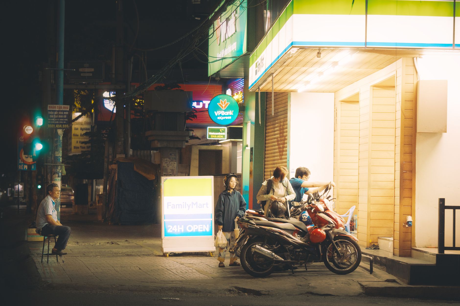 Street scene at night featuring people, motor scooters, and a brightly lit store entrance.
