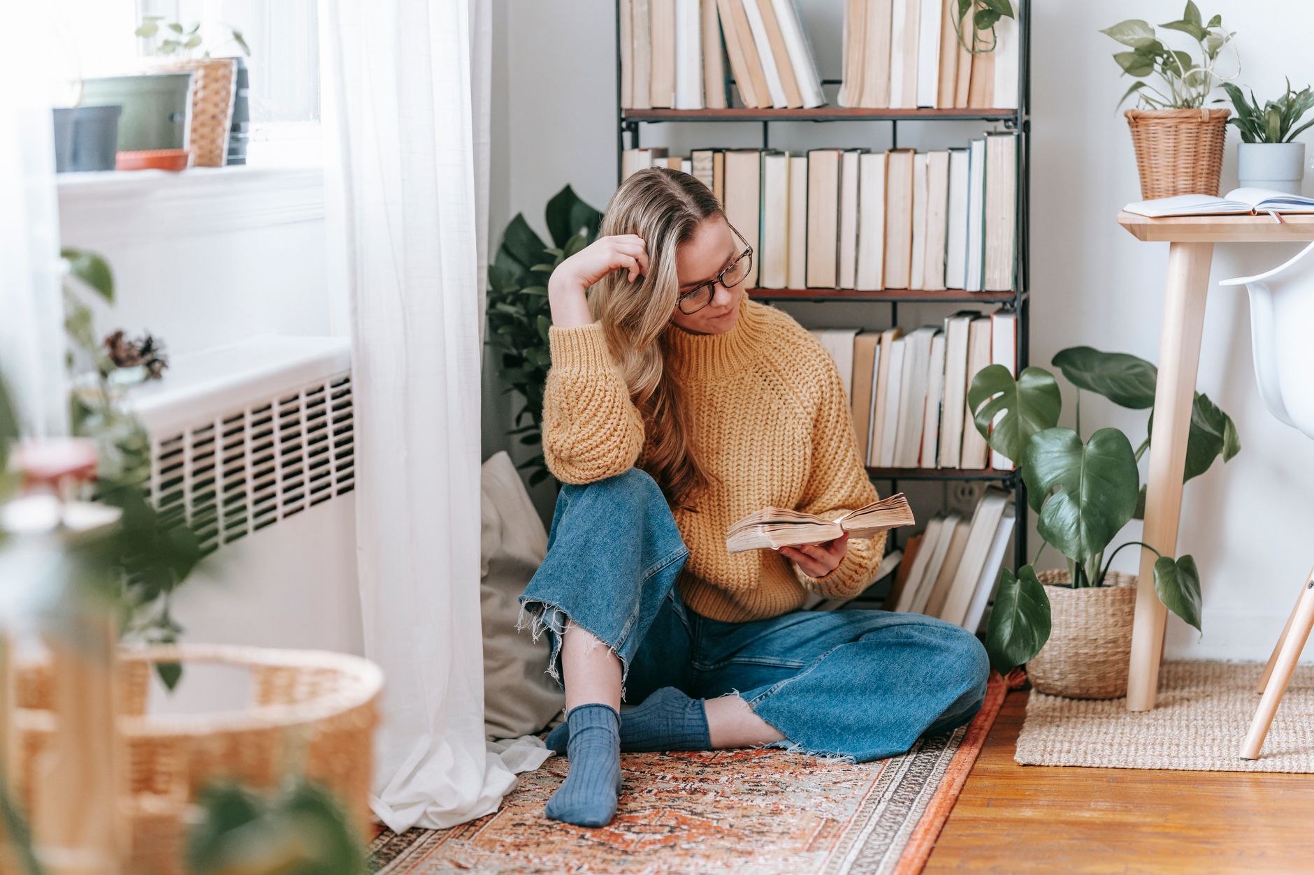 Woman reading in a cozy corner with plants and books, wearing a knitted sweater.