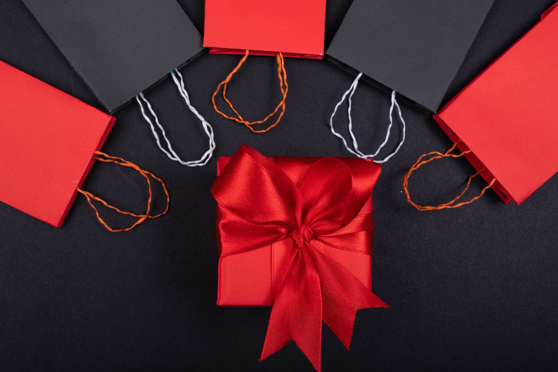 Red gift box with a bow surrounded by shopping bags on a black surface.