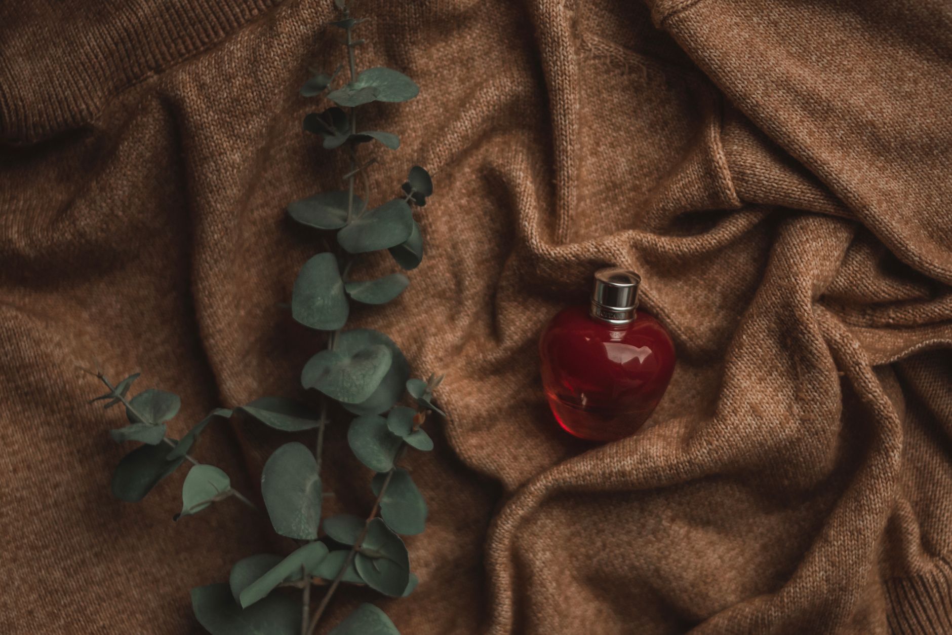 Luxurious red perfume bottle styled on cozy brown fabric with green eucalyptus leaves for a chic aesthetic.