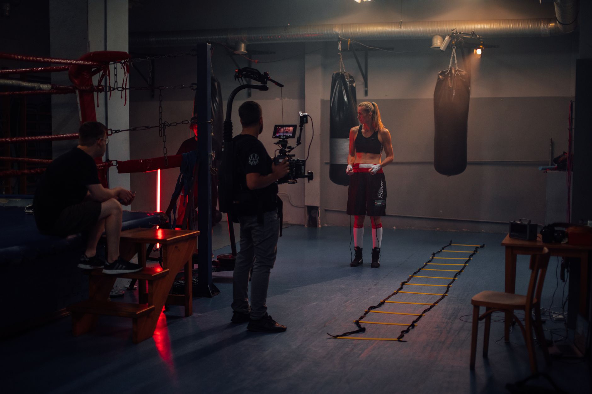 Female boxer preparing in a dimly lit boxing gym with a film crew.