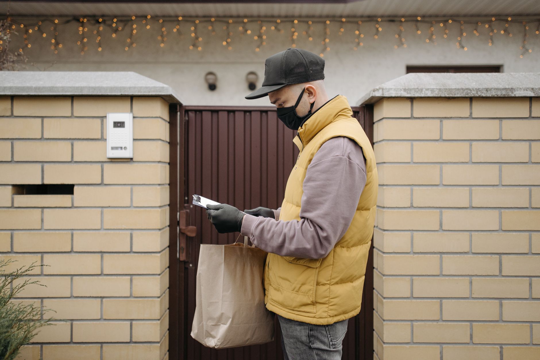 Delivery person wearing mask and gloves, holding a paper bag by a brick wall and gate.