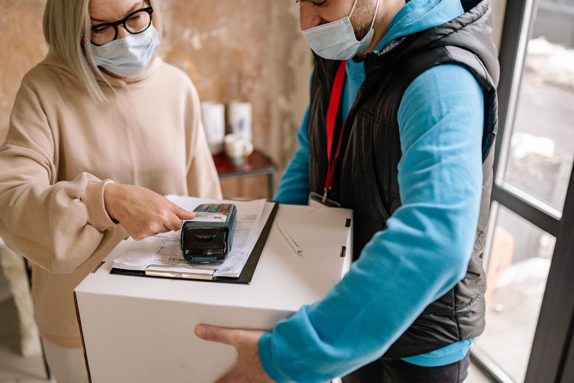 A woman makes a contactless payment for a delivery with safety measures like face masks.