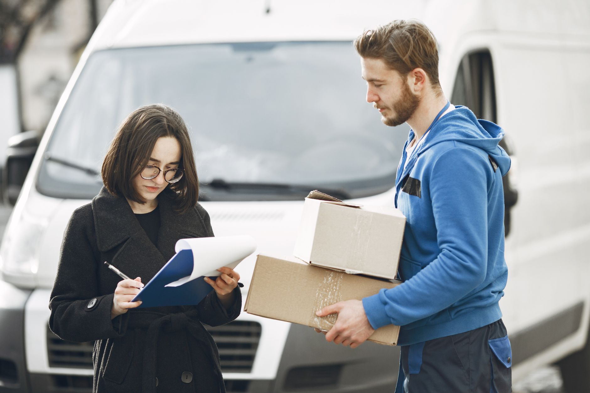 A courier in blue hands packages to a woman signing documents in front of a delivery van.