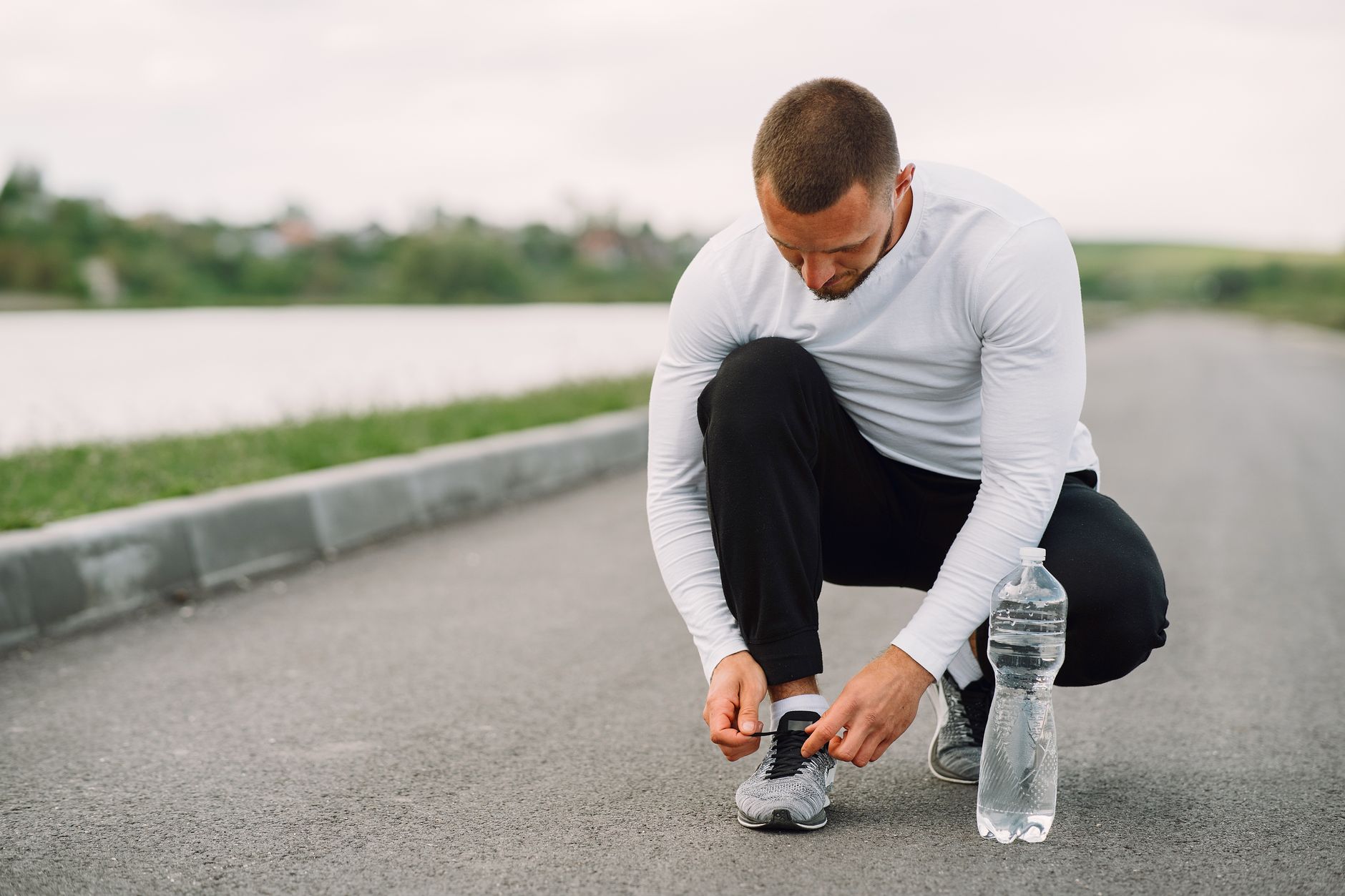 Man tying his running shoes on a rural road, with water bottle nearby.
