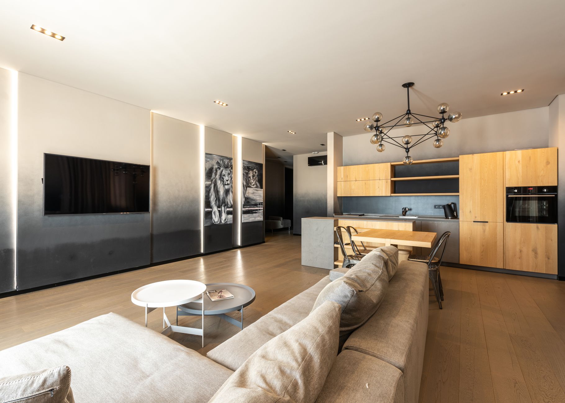 Comfortable couch placed against wall with TV placed in modern spacious apartment with kitchen zone with wooden cupboards and chandelier